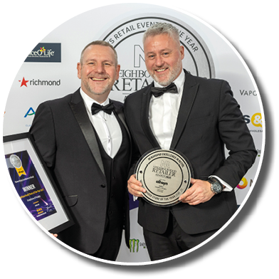 Two adults dressed in formal black tuxedos smile while holding awards at a retail excellence event. Description generated by AI