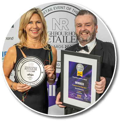 Two adults dressed in formal attire smile while holding awards at a retail event. Description generated by AI