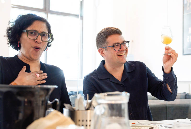 Two adults wearing glasses are sitting at a table with various items, one smiling and holding up a glass with a yellow drink while the other looks surprised with a hand on their chest. Description generated by AI
