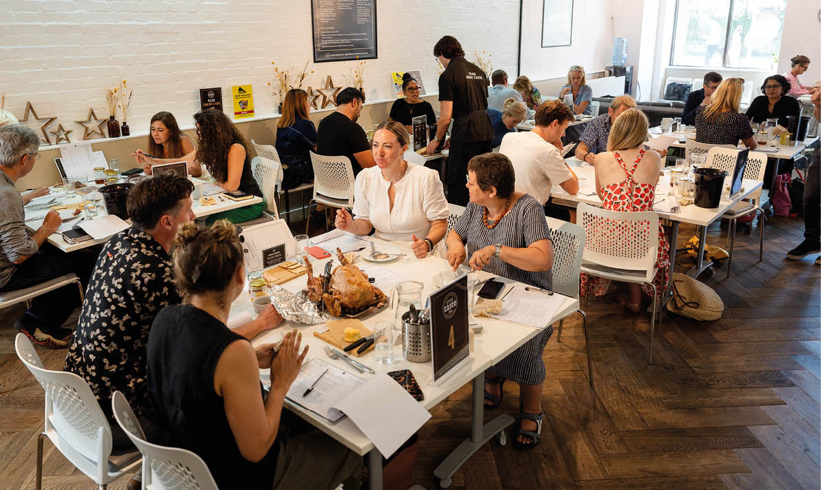 Adults sit around tables in a bright room engaged in conversation and paperwork with a roasted turkey on one table. Description generated by AI