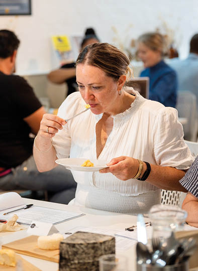 A person wearing a white blouse is sitting at a table, tasting food from a fork while holding a plate, with other people and papers visible in the background. Description generated by AI