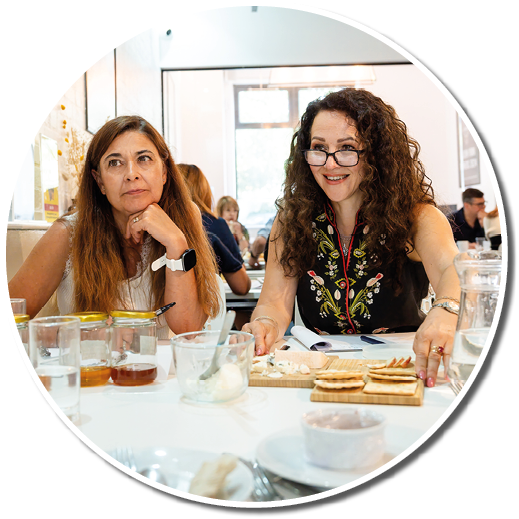 Two adults sit at a table with glasses, jars, and a wooden board with crackers and cheese, one looking thoughtful and the other smiling while wearing glasses and a floral top. Description generated by AI