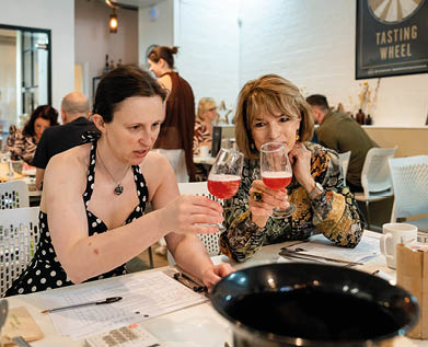 Two adults sitting at a table with papers and pens, holding glasses with a red beverage, in a casual indoor setting with other people in the background. Description generated by AI