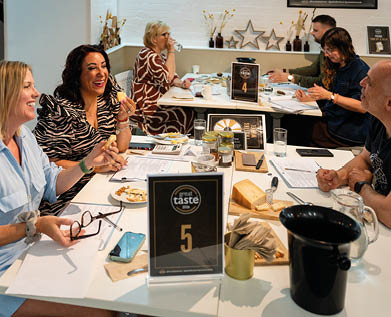 People sitting around tables in a casual setting, smiling and engaging in conversation while sampling food and drinks with papers and phones on the tables. Description generated by AI