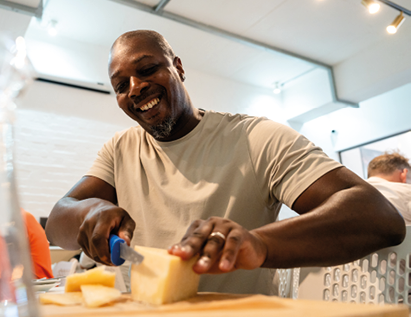 A person wearing a light-colored shirt is smiling while cutting a block of cheese on a wooden board in a bright indoor setting. Description generated by AI