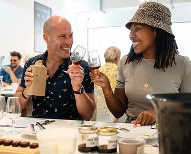 Two adults smiling and clinking wine glasses while sitting at a table with food and drinks in a bright, casual dining setting. Description generated by AI