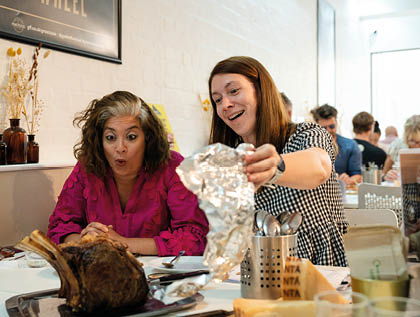 Two adults at a restaurant table excitedly uncover a large cooked meat dish while other diners are visible in the background. Description generated by AI