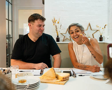 Two adults sit at a table with cheese and plates while one person smiles and holds a piece of cheese near their mouth and the other looks on with a slight smile. Description generated by AI