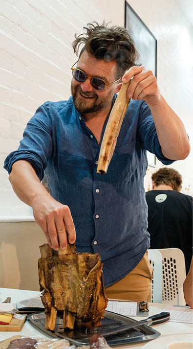 A person wearing sunglasses and a blue shirt is holding a large piece of cooked meat with bones while smiling in a bright indoor setting. Description generated by AI