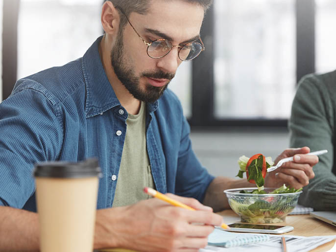 A person wearing glasses and a blue shirt is writing in a notebook while sitting at a table with a salad bowl and a coffee cup. Description generated by AI
