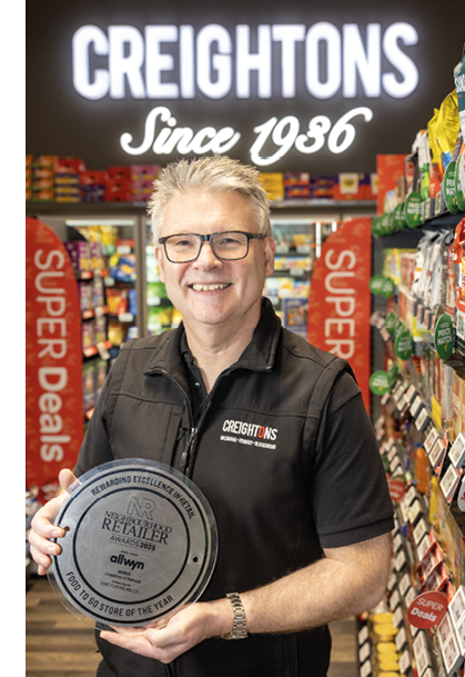 A person wearing glasses and a black Creightons shirt is smiling and holding a round award plaque inside a store aisle with shelves of products and signs reading “SUPER Deals" and "CREIGHTONS Since 1936" in the background. Description generated by AI