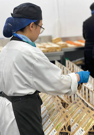 A person wearing a white coat, black apron, and blue gloves is arranging packaged sandwiches on a display shelf in a food preparation or retail setting. Description generated by AI