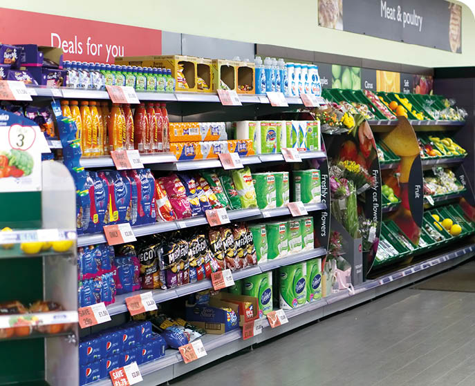 A supermarket aisle displays shelves stocked with various packaged snacks, drinks, and fresh produce under signs indicating deals and sections for fruit and vegetables. Description generated by AI