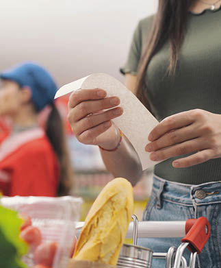 A person wearing a green shirt and jeans is holding a receipt above a shopping cart containing a baguette and other groceries. Description generated by AI