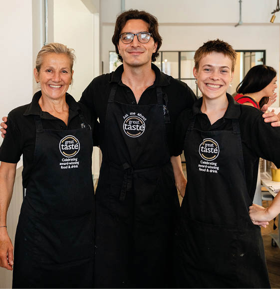 Three adults wearing black aprons with “great taste" logos smile and pose together in a bright indoor setting. Description generated by AI