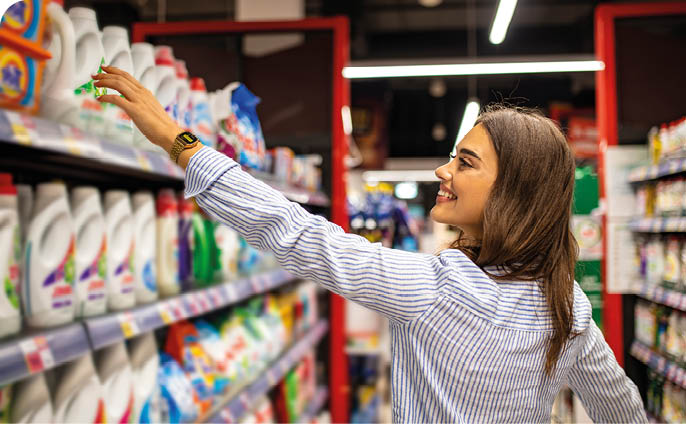 A person with long hair wearing a striped shirt is reaching for a product on a shelf in a brightly lit store aisle. Description generated by AI