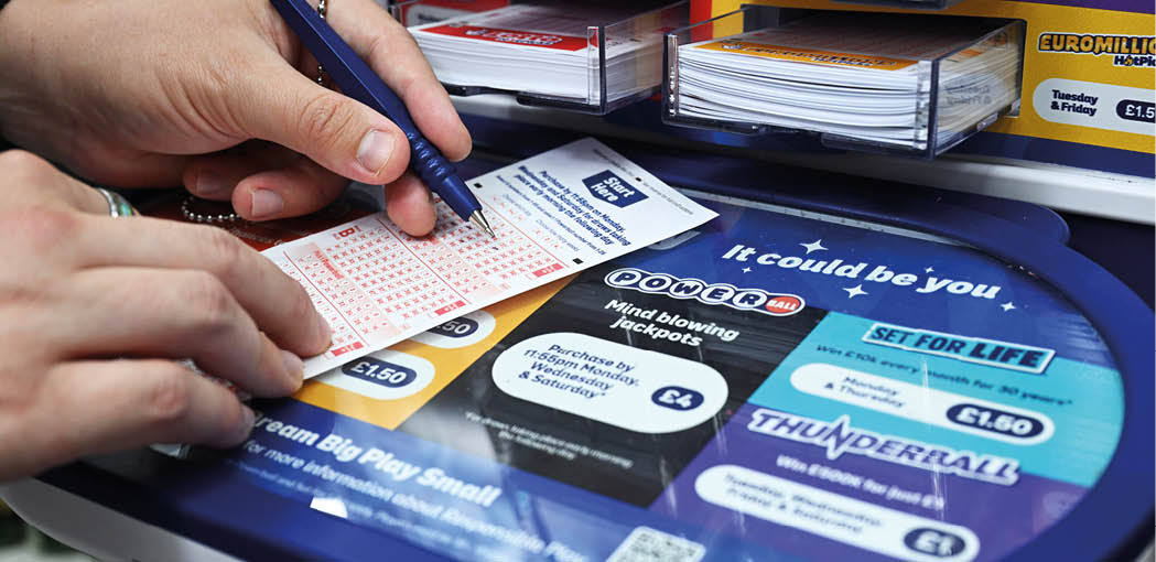 Hands are holding and marking a lottery ticket on a counter displaying various lottery game options and prices. Description generated by AI