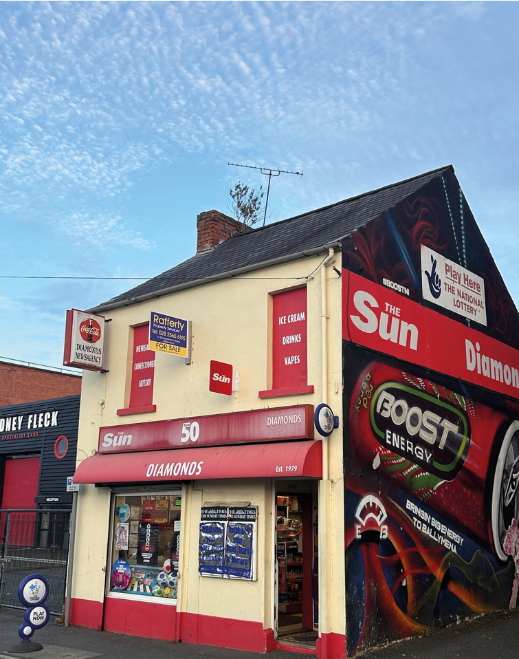 A small corner shop with red and white signage advertising The Sun newspaper, diamonds, ice cream, drinks, and wraps, located on a street with a clear blue sky above. Description generated by AI