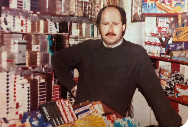 A person with a mustache wearing a dark sweater stands behind a counter filled with various packaged goods in a store. Description generated by AI
