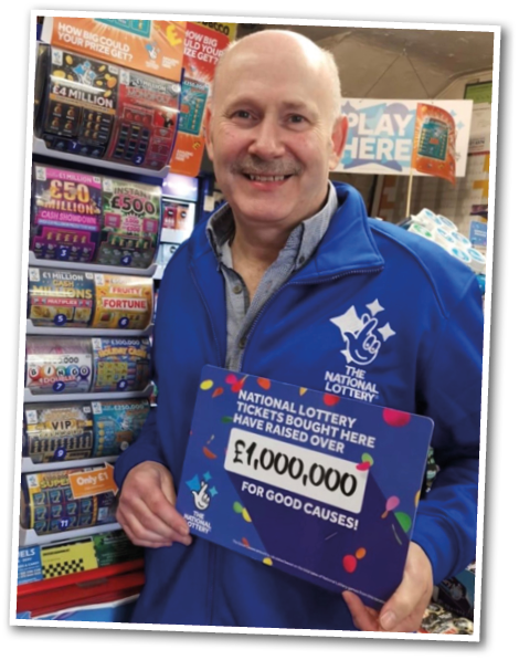 A smiling adult wearing a blue National Lottery jacket holds a sign announcing that National Lottery tickets bought there have raised over one million pounds for good causes, standing in front of a display of lottery tickets. Description generated by AI
