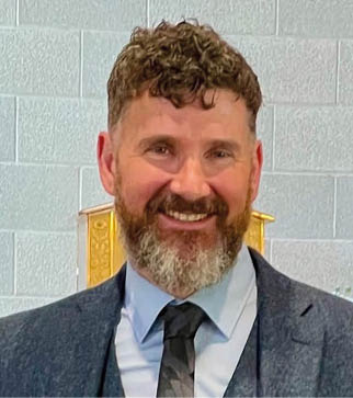 A person with curly hair and a beard is smiling and wearing a suit with a light blue shirt and dark tie against a light-colored brick wall background. Description generated by AI