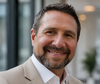 A person with short dark hair and a beard is smiling warmly while wearing a light-colored suit jacket and white shirt in an indoor setting. Description generated by AI