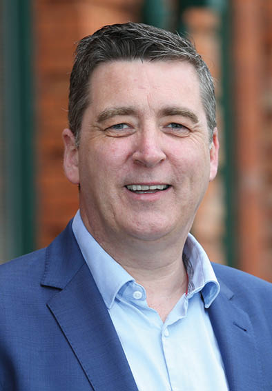 A person with short dark hair wearing a blue blazer and light blue shirt is smiling in an outdoor setting with a blurred brick background. Description generated by AI