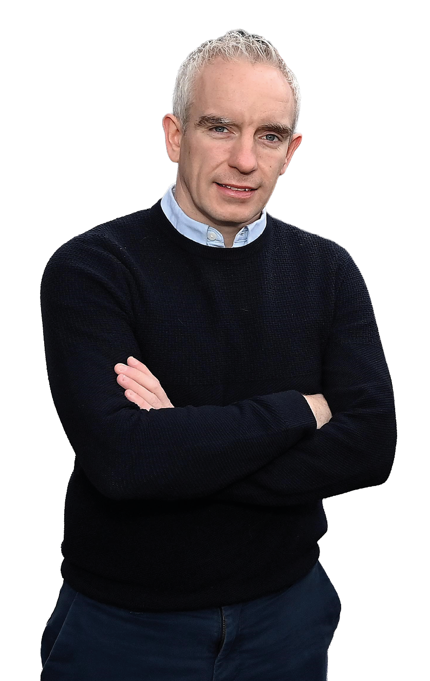 An adult with short gray hair wearing a dark sweater over a light collared shirt stands with arms crossed against a white background. Description generated by AI