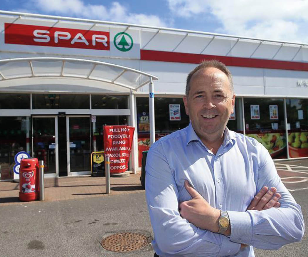 A person wearing a light blue shirt stands with arms crossed and smiles in front of a SPAR grocery store on a sunny day. Description generated by AI