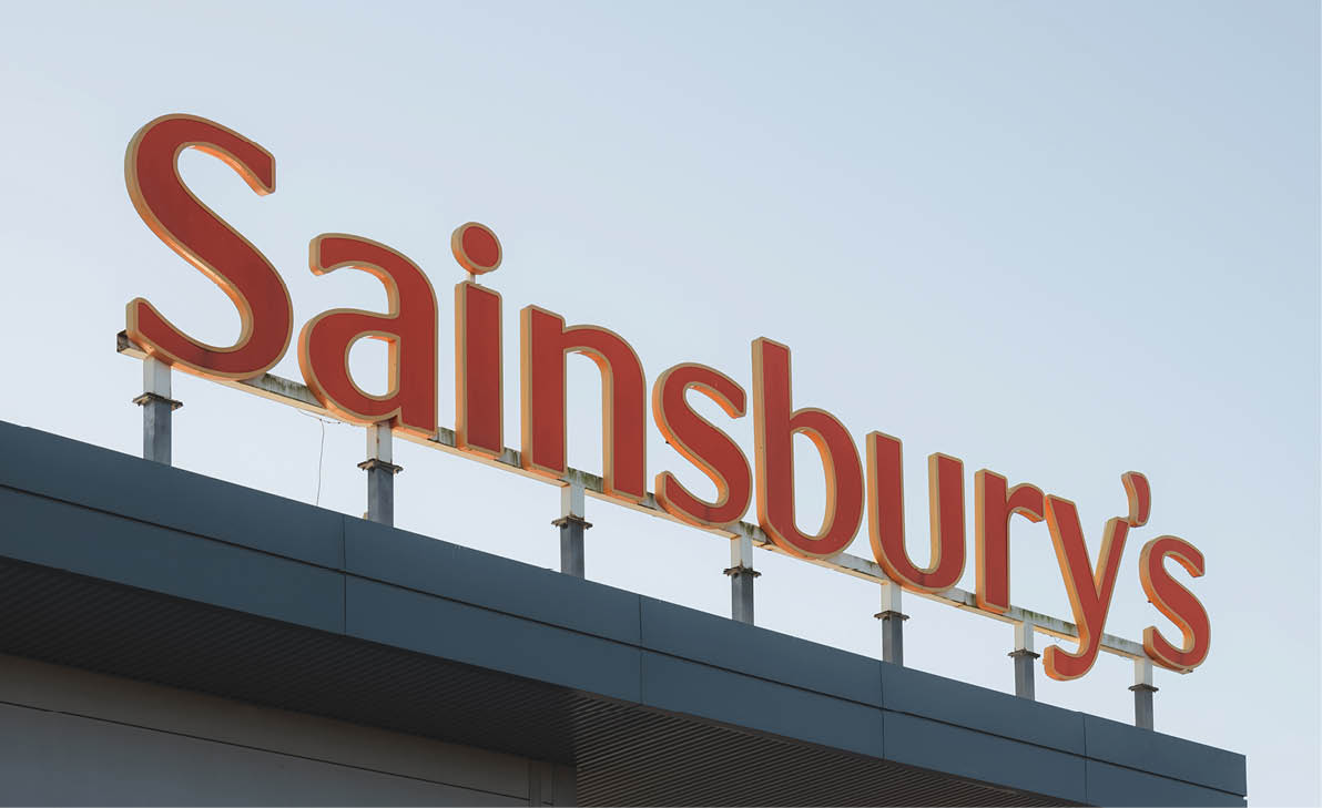 The large red and orange Sainsbury's sign is mounted on the roof of a building against a clear sky. Description generated by AI