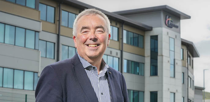A smiling senior wearing a striped shirt and dark blazer stands outdoors in front of a modern office building. Description generated by AI
