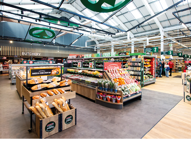 A brightly lit supermarket interior with neatly arranged fresh produce, bread, and other food items on display shelves and several people shopping in the background. Description generated by AI