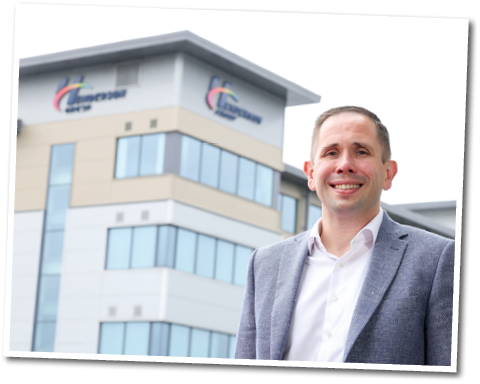 A person wearing a light-colored shirt and gray blazer is smiling and standing outdoors in front of a modern multi-story building with large windows and a company logo. Description generated by AI