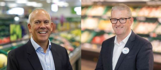 Two adults wearing business attire are smiling and standing in a grocery store produce section. Description generated by AI