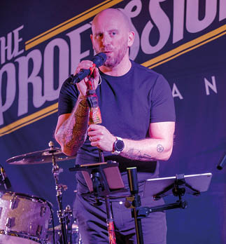 An adult with a shaved head and tattoos is singing into a microphone on stage while another adult plays an electric guitar. Description generated by AI