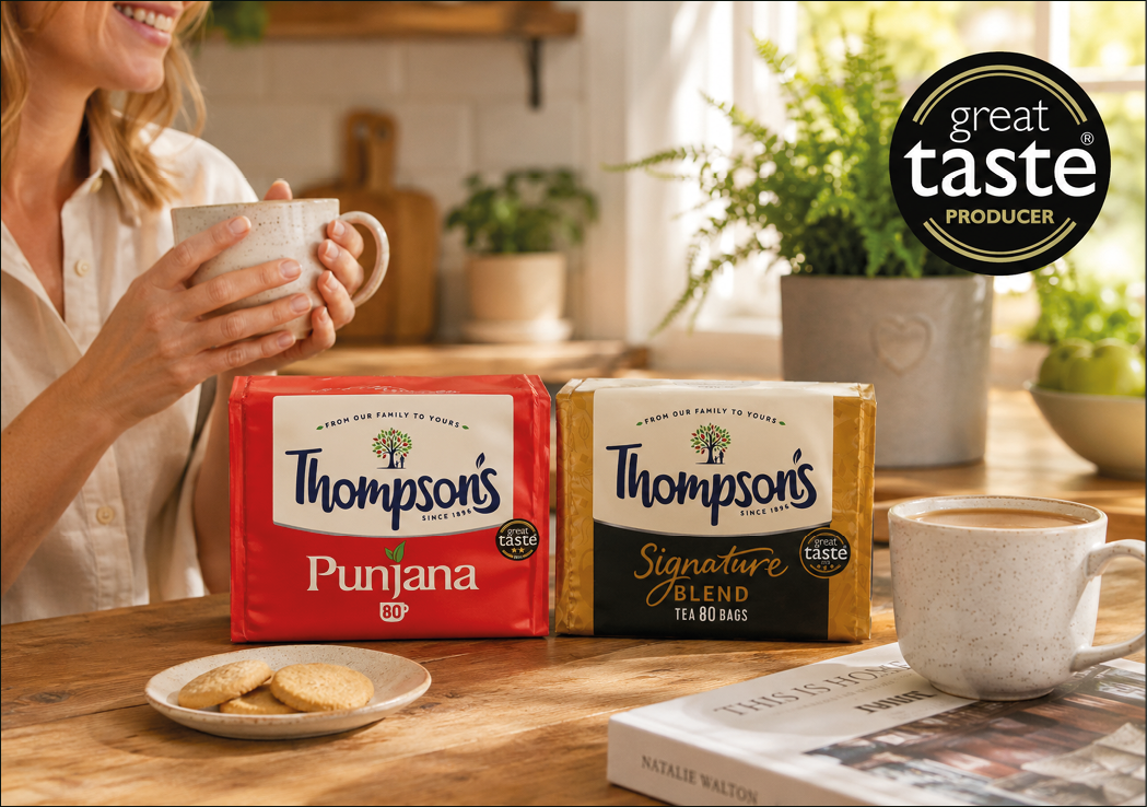 An adult holding a mug sits at a wooden table with two packages of Thompson's tea, a plate of cookies, a cup of tea, and a book in a cozy kitchen setting. Description generated by AI