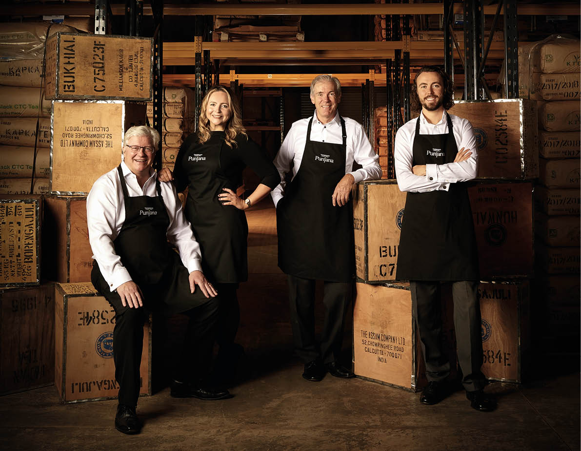 Four adults wearing black aprons stand and sit smiling in a warehouse surrounded by large wooden crates. Description generated by AI