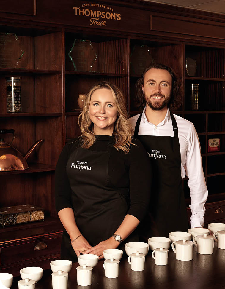 Two adults wearing black aprons stand behind a counter with multiple white cups arranged in front of them in a warmly lit wooden interior. Description generated by AI