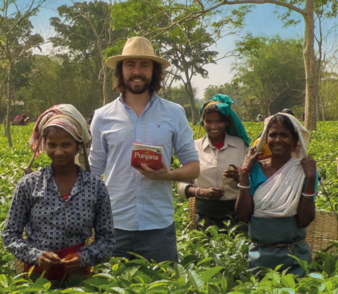 Four people standing in a green field with trees in the background, one wearing a hat and holding a red box labeled “Punjana," while the others wear headscarves and traditional clothing. Description generated by AI
