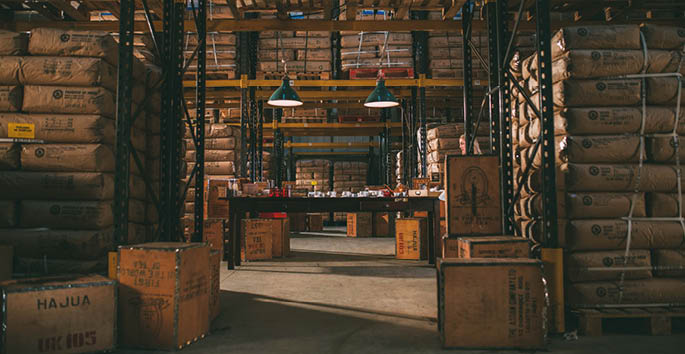 A person stands among tall stacks of large bags and wooden crates in a dimly lit warehouse with hanging industrial lights. Description generated by AI