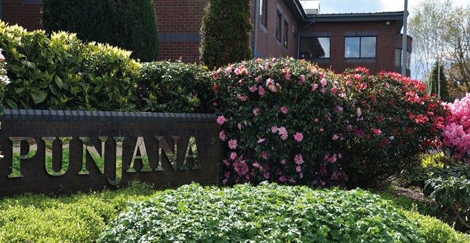 A garden area with green shrubs and pink flowers surrounds a stone sign that reads “PUNJANA" in front of a brick building under a partly cloudy sky. Description generated by AI
