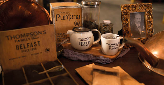 A vintage-themed display features Thompson's Family Tea products including tins, cups, and a framed black-and-white portrait on a wooden table. Description generated by AI