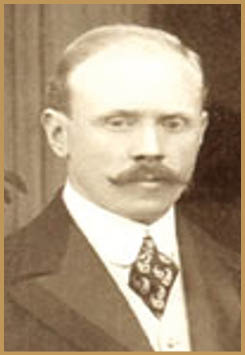 A person with a neatly combed hairstyle and a prominent mustache is dressed in a formal suit with a patterned tie and white shirt. Description generated by AI