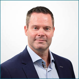 A person with short hair wearing a dark blazer and light blue collared shirt is looking directly at the camera with a neutral expression against a plain white background. Description generated by AI