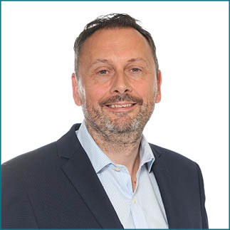 A person with short hair and a beard is smiling and wearing a dark blazer over a light blue striped shirt against a white background. Description generated by AI