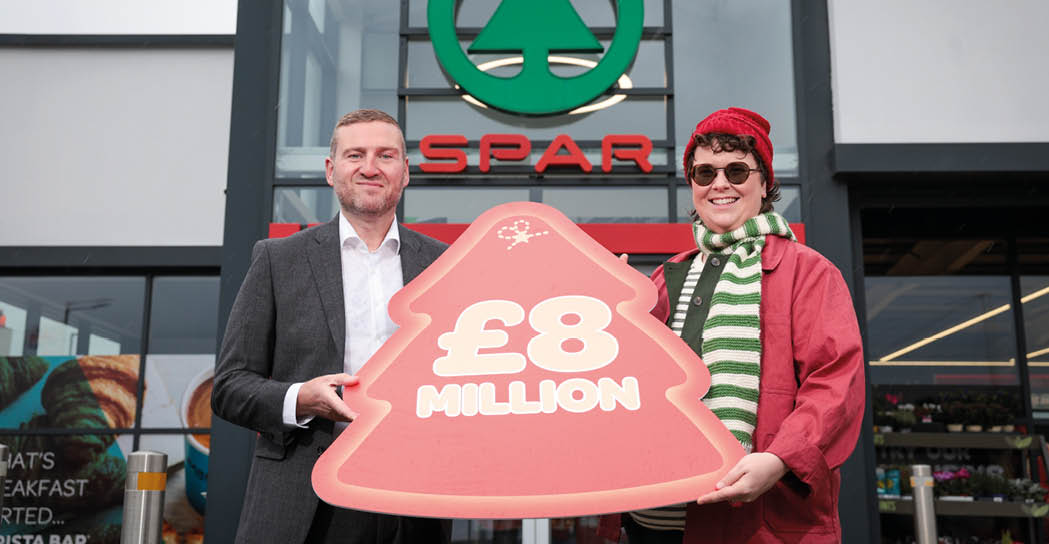 Two adults standing outside a Spar store holding a large red sign shaped like a Christmas tree that reads “£8 MILLION." Description generated by AI