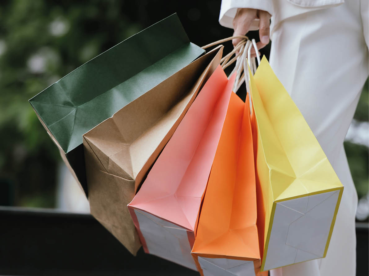 A stylish Asian woman walks happily through a modern city shopping mall. She’s holding luxury bags, enjoying the bustling urban atmosphere, surrounded by fashionable stores and other cheerful shoppers