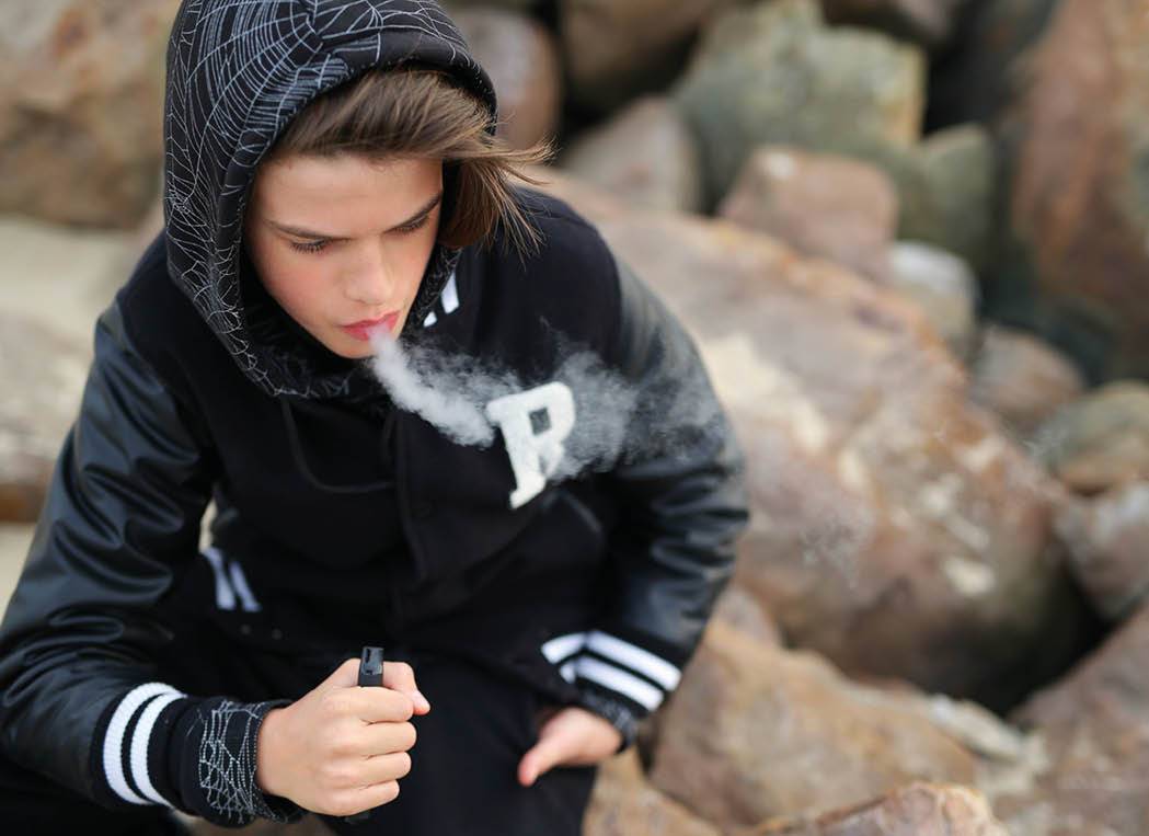 a guy of thirteen years old, holds a vape in his hands. A teenager smokes a disposable electronic cigarette while sitting by the sea