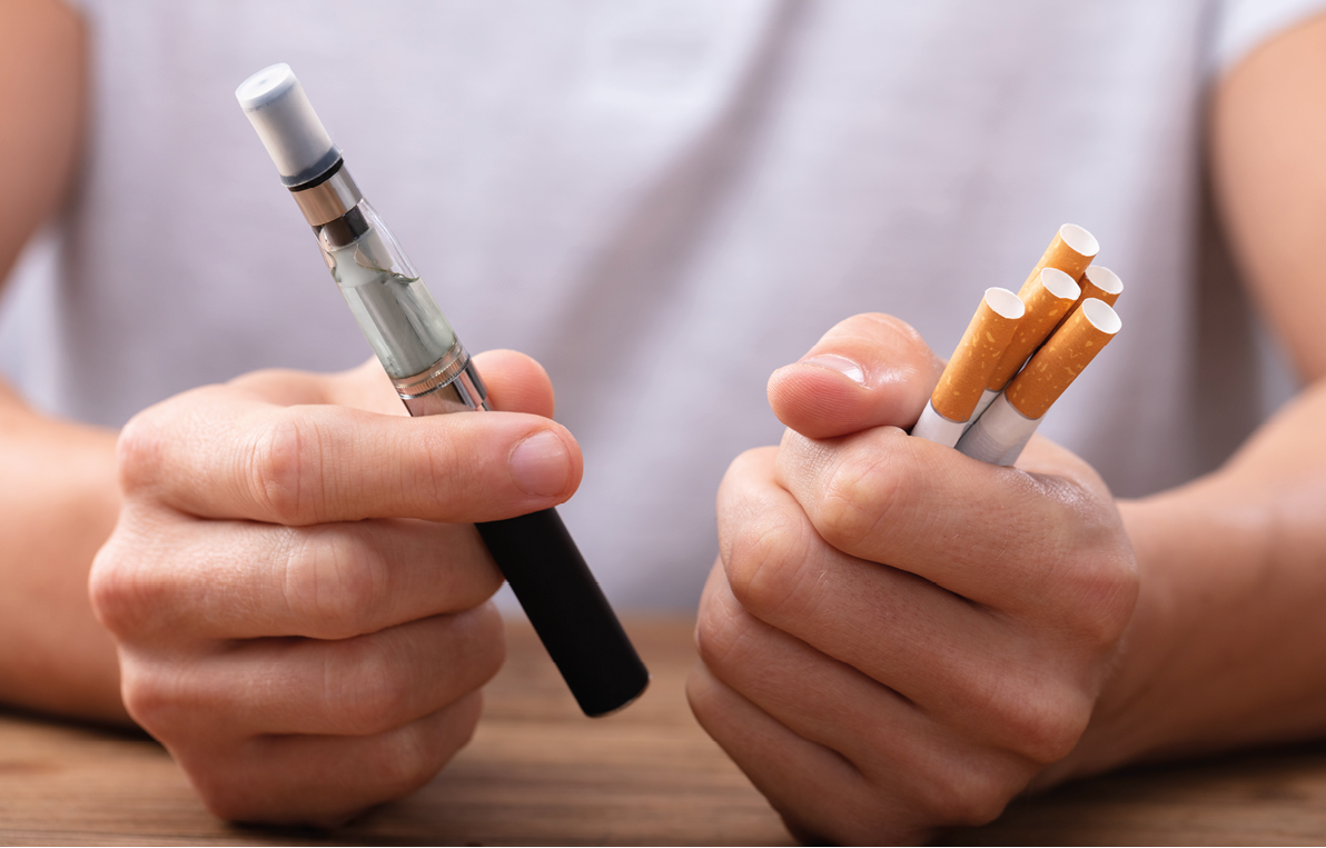 Man Holding Vape And Tobacco Cigarette Over Desk