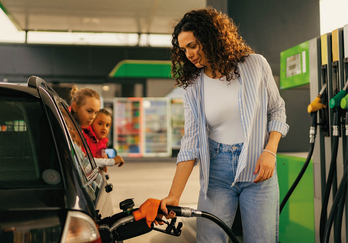 A young mother fills up gas tank at a gas station while her daughters look out the car window